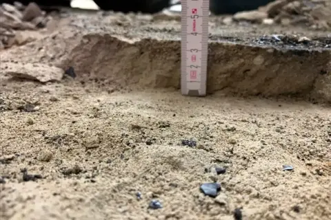 Mesure de la profondeur du décaissement du sol avec une règle. Le sable et les débris sont visibles.
