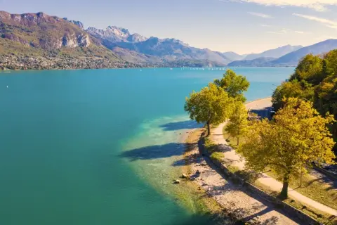 Vue aérienne du lac d'Annecy, ses eaux turquoise bordées par une route sinueuse et des arbres aux couleurs d'automne. La taille du lac est impressionnante.