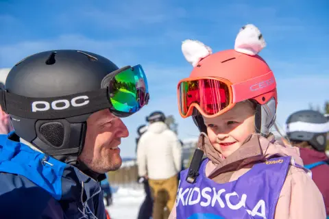 En man och ett barn i skidutrustning ler mot varandra. Barnets hjälm har söta kattöron. Perfekt för en packlista barn inför skidsemestern!