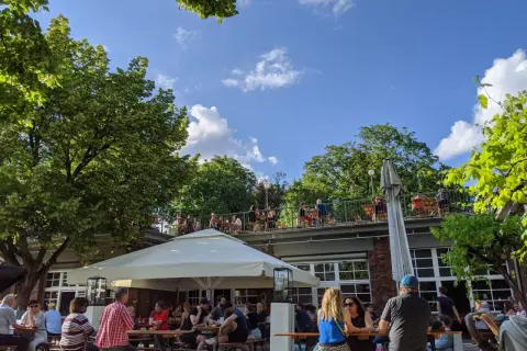 Menschen genießen das Wetter im Golgatha Biergarten. Sonnenschein, grüne Bäume und ein blauer Himmel bilden die Kulisse.