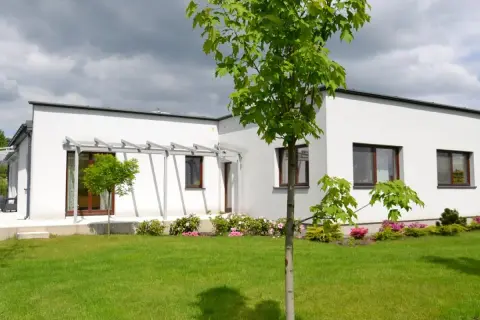 Nowoczesny bungalow z białymi ścianami i dużymi oknami, otoczony zielonym trawnikiem i kwitnącymi krzewami.