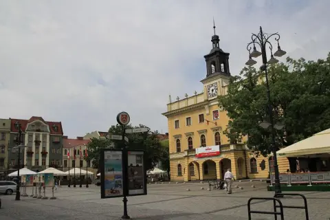 Rynek w Ostrowie Wielkopolskim z żółtym ratuszem i wieżą zegarową. W tle kamienice, drzewa i ogródek kawiarniany.