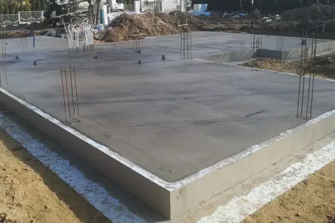 Świeżo wylany betonowy fundament pod dom, z wystającymi prętami zbrojeniowymi. To kluczowy etap, jak zrobić fundament pod dom, zapewniający stabilność konstrukcji.