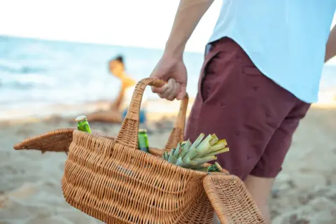 Koszyk z napojami i ananasem, idealne przekąski na plażę. W tle widać parę odpoczywającą nad morzem.