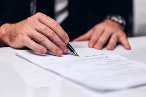 Dłoń mężczyzny w garniturze trzymająca pióro nad dokumentem.