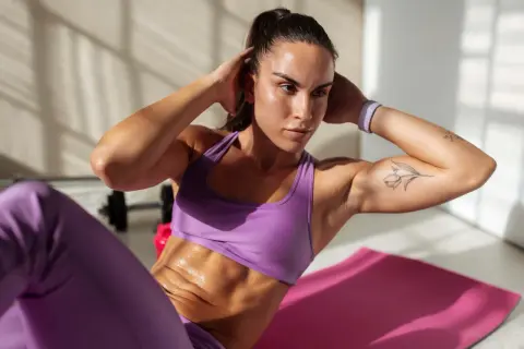 Frau macht Crunches auf einer rosa Matte, um einen flachen Bauch zu bekommen. Sie trägt lila Sportkleidung und hat ein Tattoo auf dem Arm.