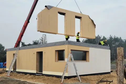 Budowa domu z gotowych elementów. Dźwig podnosi moduł dachu, pracownicy na konstrukcji montują kolejny element.