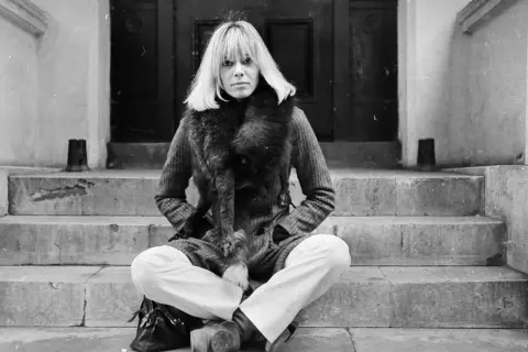 Anita Pallenberg, icona di stile, posa con un cappotto di tweed e una pelliccia voluminosa, seduta sulle scale di un portone.