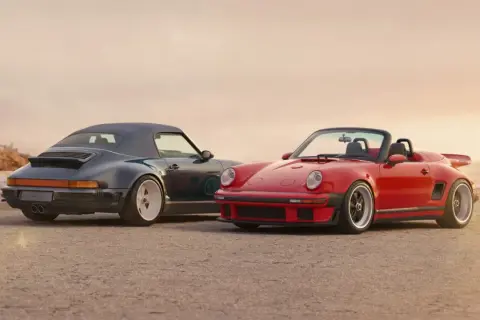 Dos Porsche descapotables, uno rojo y otro azul oscuro, posan en la playa. El **Singer Porsche** rojo destaca con su diseño deportivo y el azul con su capota puesta.