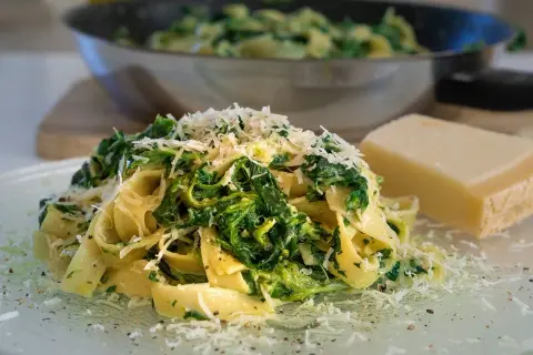 Pyszne potrawy ze szpinakiem świeżym, podane z makaronem tagliatelle i startym parmezanem. Obok leży kawałek sera.