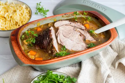 Saftiger schweinebraten im schmortopf, mit Karotten und Lauch, garniert mit Petersilie. Daneben eine Schüssel mit Spätzle.