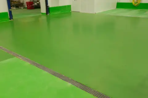 Suelo de nave industrial de color verde brillante, con un desagüe metálico en diagonal.