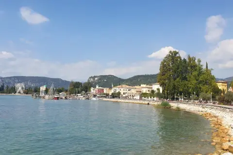 Il lungolago di Bardolino, con il lago azzurro, le barche, le case colorate e le montagne sullo sfondo. Una ruota panoramica si staglia all'orizzonte.