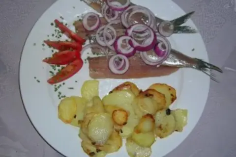 Matjes mit Bratkartoffeln, garniert mit Zwiebelringen und Tomaten. Ein klassisches Gericht, das auf einem weißen Teller serviert wird.