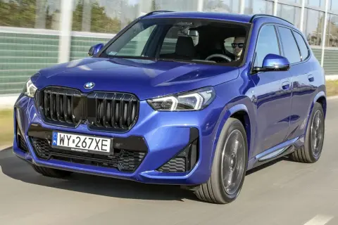 Niebieskie BMW X1 w ruchu drogowym. Kierowca w okularach przeciwsłonecznych. Zbliżenie na przód auta, widoczne logo BMW i atrapa chłodnicy. Wymiary BMW X1 pozwalają na komfortową jazdę.