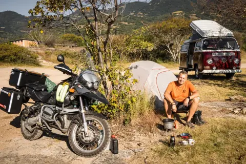 Motocykl, namiot, kamper i mężczyzna siedzący przy ognisku na tle gór.