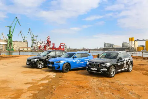 Trzy samochody BMW X: czarny, niebieski i kolejny czarny, stoją na placu budowy z dźwigami w tle.
