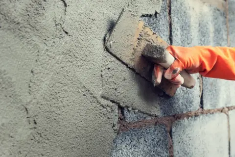 Mano con guanto arancione stende malta da costruzione su un muro di blocchi di cemento con una cazzuola.