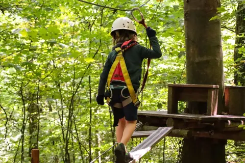 Kind im Hochseilgarten K1 balanciert auf einem schmalen Steg, gesichert durch Klettergurt und Helm.