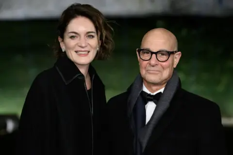 Felicity Blunt y Stanley Tucci sonríen a la cámara, vestidos elegantemente para un evento.