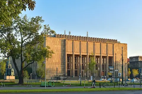 Muzeum Narodowe w Krakowie przy Alei 3 Maja. Monumentalna budowla z kamienia, otoczona zielenią i stojakami z hulajnogami.