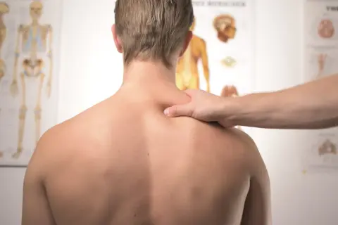 Dłoń ortopedy badająca plecy pacjenta na tle anatomicznych plakatów.