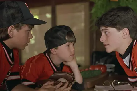 Trzej chłopcy w strojach baseballowych, przypominający scenę z filmów dla dzieci z lat 90. Jeden z nich trzyma rękawicę z piłką.