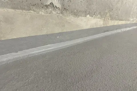 Szara posadzka z widoczną dylatacją wypełnioną białym materiałem, granicząca z betonową ścianą.