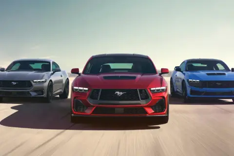 Tre Ford Mustang modeller i rött, grått och blått kör på en ökenväg.