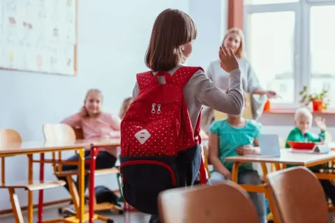 Dziewczynka z czerwonym plecakiem w klasie, może odczuwać lęki u dziecka 7 latka, ale nauczycielka i koledzy dodają jej otuchy.