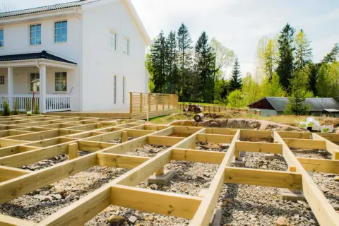 Grundläggande stomme för att bygga veranda framför ett vitt hus. Träbjälkar ligger på grus och stenar, redo för nästa steg.
