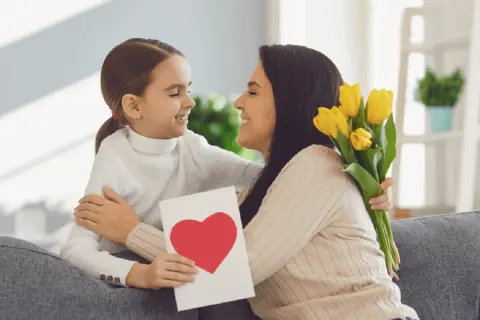 Uśmiechnięta dziewczynka wręcza mamie kartkę z sercem i bukiet żółtych tulipanów. Radosny Dzień Matki UK!