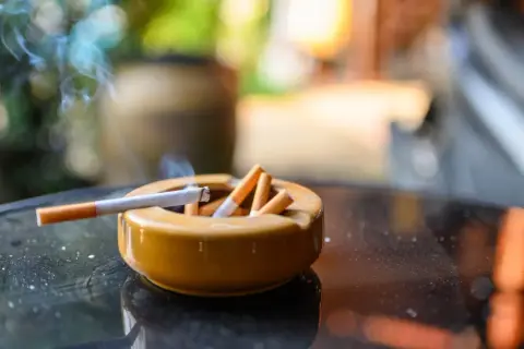 Cenicero lleno de colillas, con un cigarrillo encendido y humo. Un recordatorio de la necesidad de eliminar olor tabaco casa.