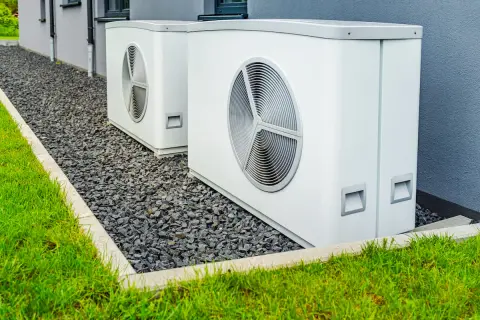 Due unità esterne di pompe di calore alta temperatura, bianche e moderne, installate su ghiaia accanto a un prato verde e a un edificio grigio.