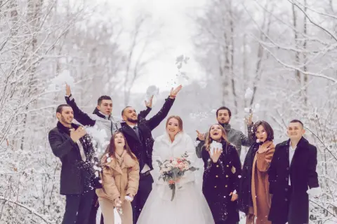 Brautpaar und Gäste werfen Schnee in der Luft. Perfekt für eine Hochzeit im Winter: Was anziehen? Warme Mäntel und festliche Kleidung.