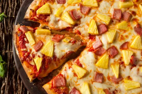 Pyszna hawaiian pizza z kawałkami ananasa i szynki, obficie posypana serem, prezentuje się apetycznie na ciemnym tle.