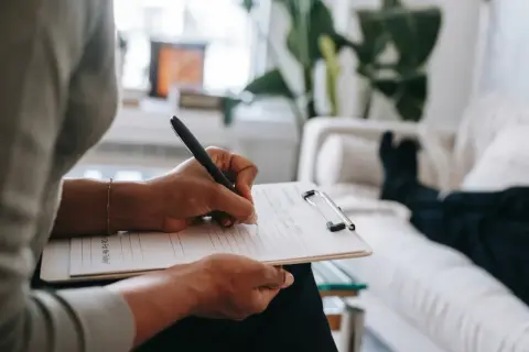En psykiater eller psykiatriker skriver anteckningar under en terapisession. En person ligger på en soffa i bakgrunden.