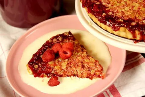 En bit smulpaj med hallon och krämig vaniljsås på en rosa tallrik. En hel smulpaj syns i bakgrunden.