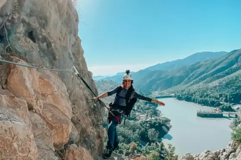 Aventura en el Caminito del Rey. Mujer sonríe mientras recorre la vía ferrata, con un embalse y montañas de fondo.