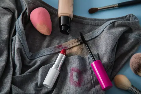 Graues T-Shirt mit Flecken von Lippenstift, Mascara und Foundation. Ein Make-up-Schwamm und Pinsel liegen daneben. Tipps zum Make-up aus Kleidung entfernen.