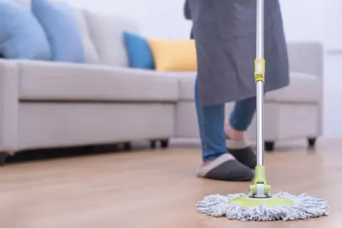 Persona con delantal y zapatillas limpiando el suelo con una mopa. Aprende como limpiar la mopa después de usarla para mantener tu hogar impecable.