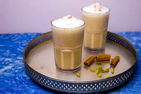 Två glas med krämig chai latte, toppad med skum och kanel. Kardemumma och kanelstänger på brickan ger en hint om detta chai latte recept.