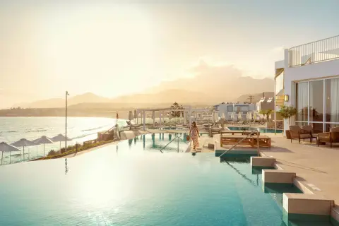 En kvinna i en blommig klänning går mot en infinitypool vid havet på Kreta, Makrigialos. Solnedgången färgar himlen gyllene.