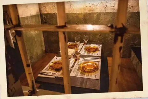 Mesa puesta para el señor cachopo, con platos ámbar y copas, tras unos barrotes.