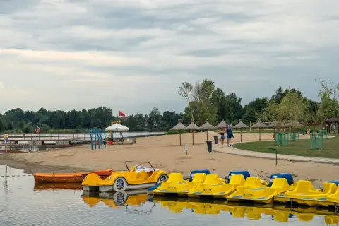 Żółte rowerki wodne i łódki czekają na plaży nad jeziorem blisko Warszawy. Para spaceruje po piasku pod parasolami.