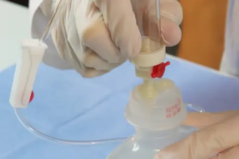 Ręce w rękawiczkach przygotowują kroplówkę, łącząc dren z pojemnikiem. Instrukcja jak podłączyć kroplówkę.