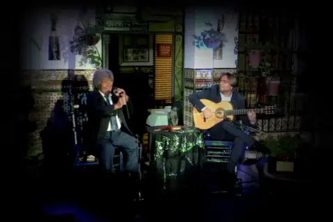Dos hombres disfrutan de un emotivo canto flamenco. Uno canta al micrófono, el otro acompaña con su guitarra.