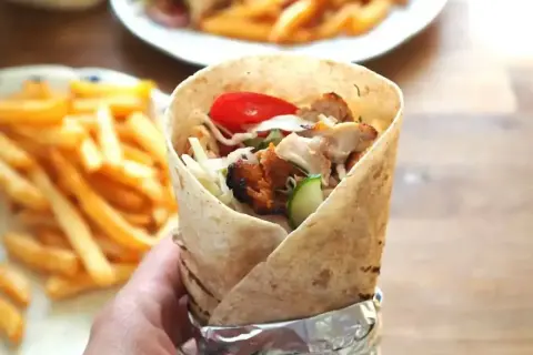 Ręka trzymająca prawdziwy kebab z kurczakiem, warzywami i frytkami w tle.