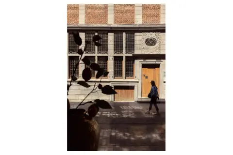 Photographie architecture : une femme passe devant une façade historique aux fenêtres multiples et à la porte en bois.