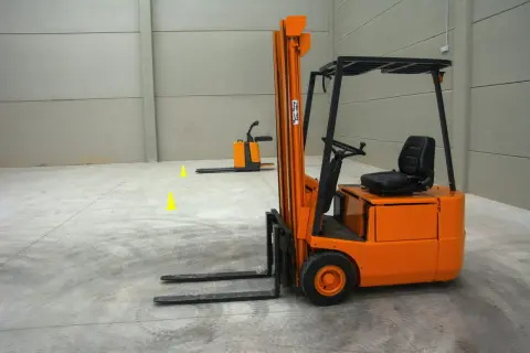 Pomarańczowy wózek widłowy z szerokim rozstawem wideł, gotowy do pracy. W tle inny wózek i żółte stożki.